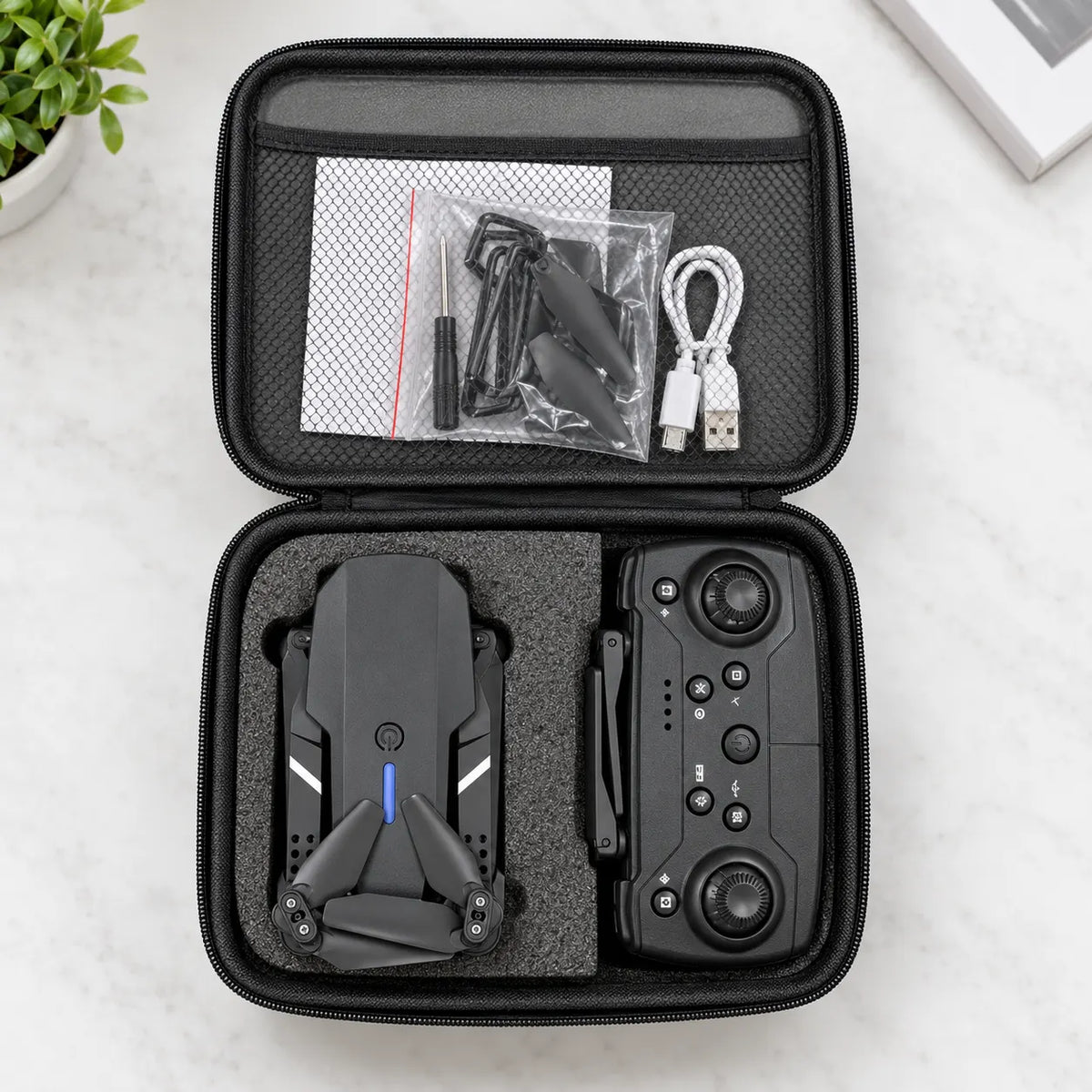 An open black hard carrying case laid on a light marble surface, showing a folded black drone inside the left compartment and a black remote controller in the right. The case interior also holds a USB cable, small accessories in a zip bag, and a white pouch.